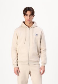 Beigā krāsā zip-up kapuce ar regulējamu kapuci un priekš�ējām kabatām. Ar zilu logo izšuvumu uz krūšu daļas. Mīksta kokvilnas auduma.