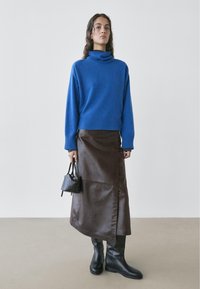 Woman wearing a blue turtleneck sweater, dark brown leather midi skirt, black knee-high boots, and holding a small black handbag.