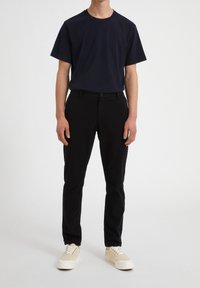 Man som bär svarta slim fit byxor, marinblå kortärmad t-shirt och beige sneakers, stående mot en enkel ljus bakgrund.
