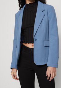 Svetle modrý blejzr s přizpůsobeným designem, s klopami, zapínáním na jeden knoflík a hladkým povrchem. Nosený přes černý žebrovaný top.