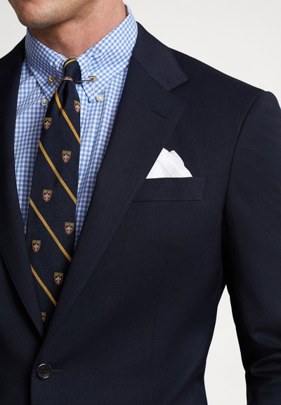 Tamsiai mėlynas švarkelis su aptemptu dizainu, derinamas su šviesiai mėlyna languota marškinėlių ir dryžuota kaklaraiščiu, papuoštu herbo logotipais. Baltas kišeninis lapelis kaip akcentas.