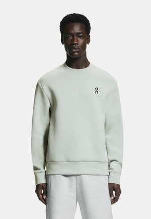 Férfi világoszöld kereknyakú pulóverben, amelyen egy kis fekete logó található a mellkasán, világosszürke melegítőnadrágban, fehér háttér előtt áll.