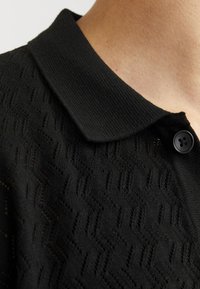 Polo shirt nero in maglia con un motivo texturizzato, dotato di colletto piatto e chiusura a un bottone. Tessuto morbido con dettagli sottili.