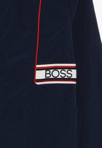 Tejido azul marino con un bolsillo diagonal contorneado en ribete rojo, que presenta una etiqueta blanca con franjas rojas y negras y el texto "BOSS."