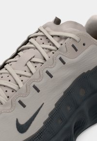 Sapatilha Nike cinza com um superior texturizado, acentos escuros contrastantes e atacadores redondos. Apresenta perfurações para ventilação e um design ondulado.