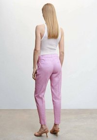 Frau mit glattem blonden Haar trägt ein weißes ärmelloses Oberteil, pinke tapered Pants und beigefarbene hohe Sandalen, steht auf einem neutralen Boden und gegen eine neutrale Wand.