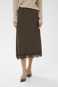 Femme portant une jupe midi marron foncé avec un ourlet en dentelle, un pull en maille beige et des escarpins marron à bouts pointus sur un fond blanc.