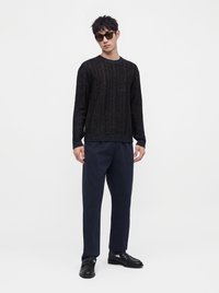 Jeune homme portant des lunettes de soleil foncées, un pull noir texturé, un pantalon bleu marine et des chaussures noires, debout devant un fond clair uni.