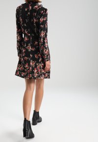 Vestido negro de flores con mangas largas, cintura ajustada y falda acampanada. Presenta patrones florales coloridos y se lleva con botas negras hasta el tobillo.