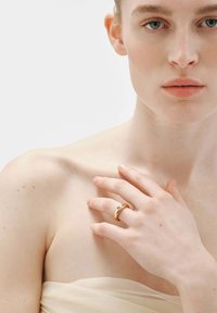 Anillo de oro con diseño curvado, que presenta una piedra preciosa clara. El modelo muestra el anillo sobre piel clara contra un fondo minimalista.