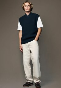 Marine gebreide mouwloze trui over een wit T-shirt, gecombineerd met crèmekleurige pantalons en zwarte schoenen met bruine accenten.