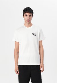 White cotton T-shirt with short sleeves, round neck, featuring a black printed logo on the left chest and a small embroidered accent on the sleeve.