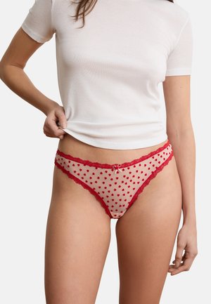 Frau hebt weißes Hemd an, trägt transparente rote Unterwäsche mit kleinen Herzmustern und Spitzenbesatz vor einfachem Hintergrund.