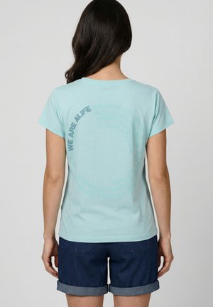 Frau mit langen dunklen Haaren, die ein hellblaues T-Shirt mit kreisförmigem Text und dunkelblaue hochgekrempelte Shorts trägt, und von der Kamera wegschaut.