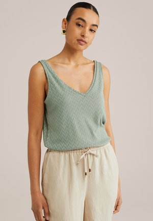 Débardeur vert clair avec un col en V et motif en chevron texturé. Associé à un pantalon beige avec cordon de serrage et accessoires décoratifs.
