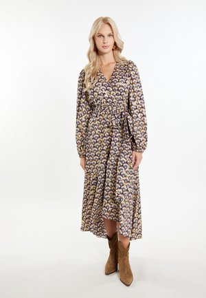 Blumengemustertes, langärmeliges Wickelkleid in Marineblau, Gold und Creme; mit einem Bindegürtel, fließendem Rock und strukturierter Stoffqualität. Braune Knöchelstiefel.