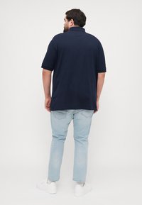 Marineblauwe polo met korte mouwen, voorzien van een klassieke kraag. Gecombineerd met lichtblauwe jeans en witte sneakers, gezien vanuit de achterkant.