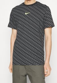 Černé tričko s krátkým rukávem s opakovaným šedým a bílým vzorem vlnky, s malým jasně žlutým logem Nike na hrudi.