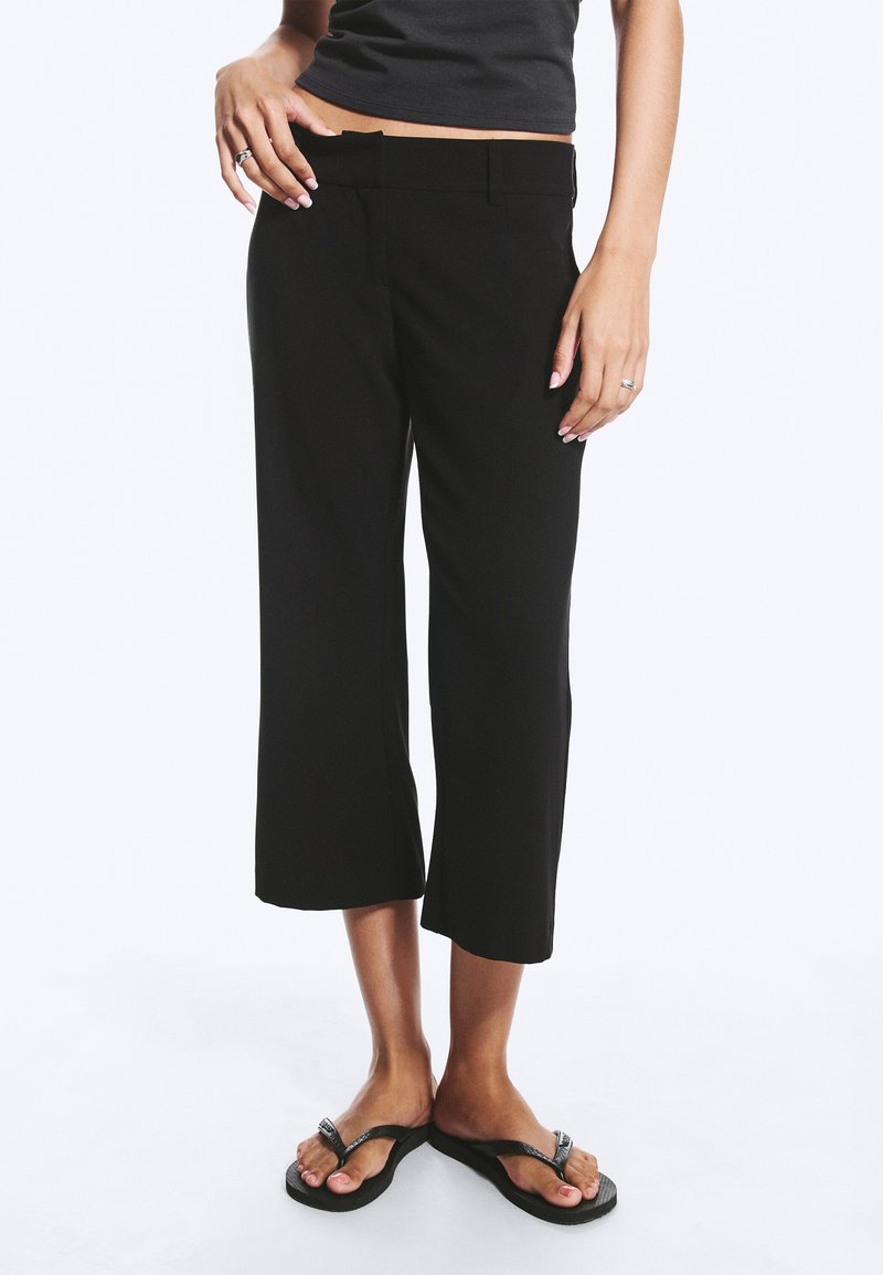 Femme portant un pantalon large court noir, un débardeur noir et des tongs noires, debout sur fond blanc.