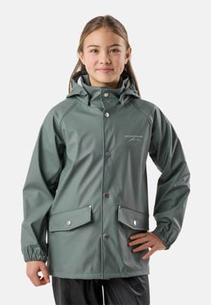 Ung person iført grøn vandtæt hættejakke med trykknaplommer, stående med den ene hånd på hoften mod en ensfarvet baggrund.