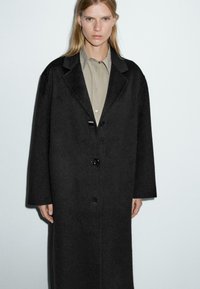 Cappotto di lana nera con una vestibilità rilassata, rever a punta e tre bottoni. Indossato sopra una camicia chiara con colletto. Texture liscia.