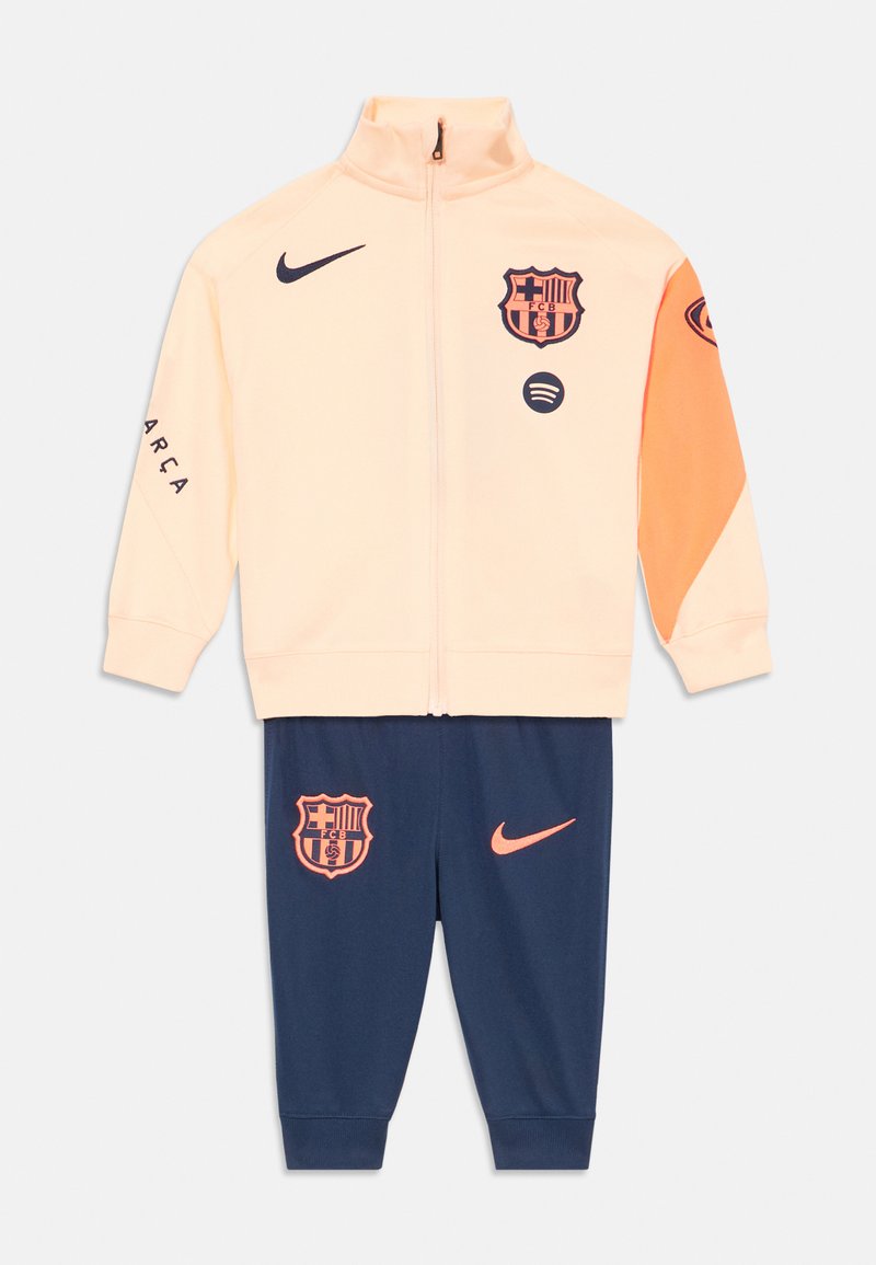 Barcelona-teemainen treenipuku, jossa on vaalean persikan värinen vetoketjuhuppari oransseilla yksityiskohdilla ja tummansiniset housut, joissa on logoja ja vaakunoita.