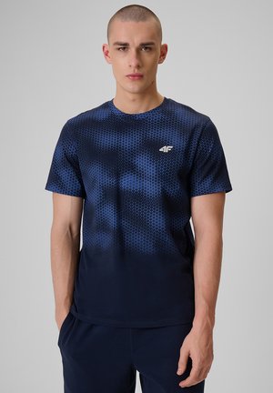 Młody mężczyzna z krótkimi włosami, ubrany w granatowy T-shirt z geometrycznym wzorem i granatowe spodnie, stoi z rękami w kieszeniach.