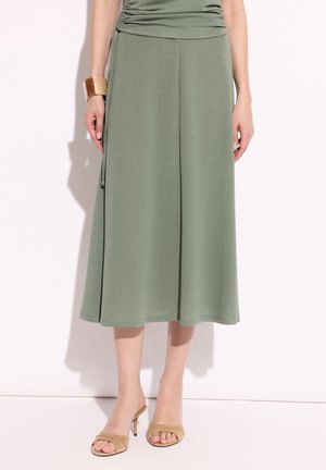 Kobieta w oliwkowej spódnicy midi, beżowych sandałach na wysokim obcasie i złotej bransoletce mankietowej, stoi na białym tle.