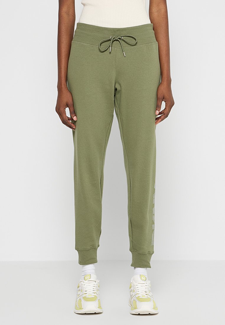 Persona che indossa pantaloni jogger verde oliva con vita regolabile, abbinati a sneaker bianche e gialle e calze bianche.