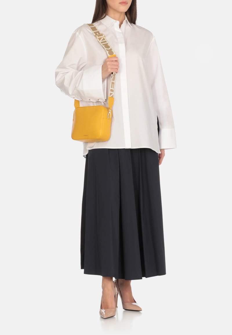 Borsa a tracolla gialla in pelle con superficie texturata, dotata di una tracolla color marrone chiaro con stampa del logo. Indossata con una camicia bianca e una gonna nera.