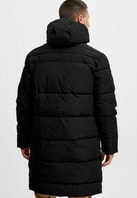 Sort pufferjakke med hætte, der har et quiltet design, lang længde og elastiske manchetter. Viser en glat, mat tekstur.