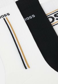 Drei Paare Socken: ein weißes mit einem schwarzen und goldenen Streifen, eines in Schwarz und ein weißes mit einem Logo. Rippstrick-Textur, eng anliegendes Design.