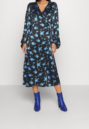 Vrouw draagt een zwarte wikkeljurk met een blauw bloemmotief en lange mouwen, gecombineerd met felblauwe enkellaarsjes, staand op een grijze vloer.