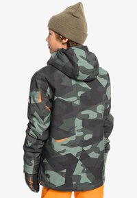 Kamouflagemönstrad jacka i mörkgrön och grå, med justerbar huva och orange detaljer. Bärs med en stickad mössa.