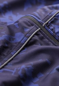 Κλειστό πλάνο σκούρου μπλε υφάσματος με μαύρο φερμουάρ και διακριτικό μπλε καμουφλάζ, με ανάκλαστη ταινία κατά μήκος των ραφών.