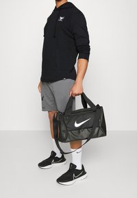 Černá sportovní taška Nike s bílým logem, odolná tkanina, dvojité držadla na nošení a zipové uzavření. Model v šedých šortkách a černých teniskách.