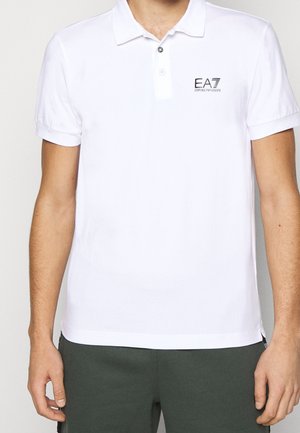 Mężczyzna ubrany w białą koszulkę polo EA7 Emporio Armani i ciemnozielone spodnie, ramiona swobodnie opuszczone wzdłuż ciała, na jednolitym białym tle.