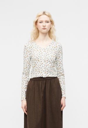 Jeune femme aux cheveux blonds portant un haut blanc à fleurs à manches longues et une jupe marron taille haute sur un fond uni.