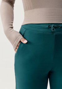 Beżowy, prążkowany top z długim rękawem w połączeniu z ciemnozielonymi spodniami z elastycznym pasem z regulowanym sznurkiem. Spodnie mają boczne kieszenie dla wygody.