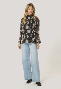 Blusa nera a fiori con maniche lunghe in organza, scollo alto e dettagli arricciati, abbinata a jeans a gamba larga di un blu chiaro e stivaletti neri.