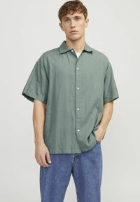 Kortærmet skjorte med knapper i dæmpet grøn stof, der har en afslappet pasform, krave og hvide knapper, parret med blå denim jeans.