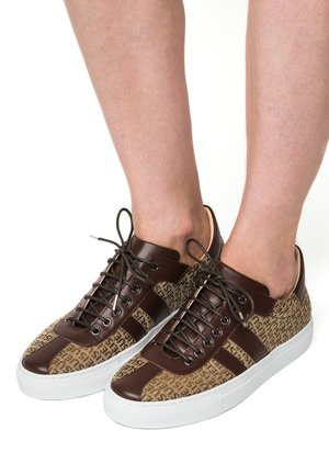 Bruine en beige low-top sneakers met donkerbruine veters, gedragen op blote voeten tegen een witte achtergrond.