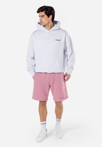 Sweatshirt branca com um bolso frontal e logótipo preto "PEGADOR", combinada com calções rosa e ténis brancos, num fundo neutro.