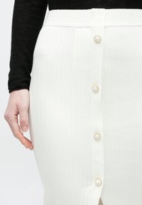 Közelkép egy kézről, amely egy fehér bordázott szoknya mellett van, aranyszegélyes gombokkal, hosszú ujjú fekete felsővel kombinálva.