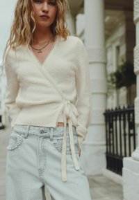 Cream knitted wrap sweater with tie detail, paired with light blue jeans featuring embellished pockets. Soft texture and relaxed fit.