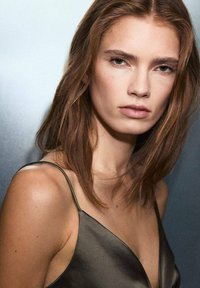 Persona con cabello castaño que lleva una camisola de satén verde oscuro con tirantes delgados, que presenta una textura suave y un corte sencillo.