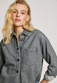 Grijze denim blouse met een knoopsluiting, een borstzak en opgerolde mouwen. Bevat zwarte knopen en contrasterende stikdetails.