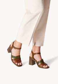 Brown and green strappy heeled sandals with a block heel, featuring horizontal and vertical straps, and an ankle buckle closure.
