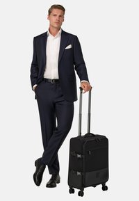 Czarna walizka na kółkach z dwoma zapięciami, teksturowanym dnem, solidnym uchwytem i gładkimi kółkami, obok mężczyzny w granatowym garniturze.