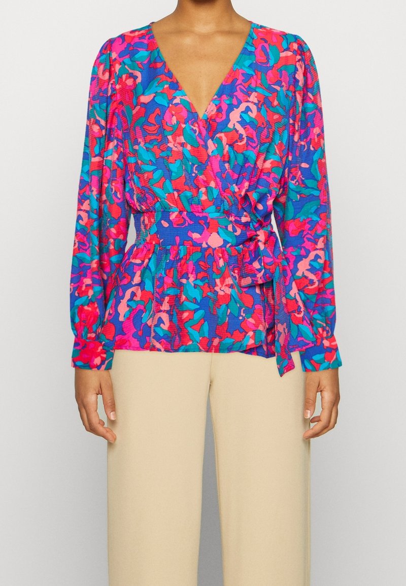 Blouse multicolore avec un col en V, des manches longues et une taille à nouer. Présente un motif abstrait vibrant dans des nuances de rose, bleu et violet.